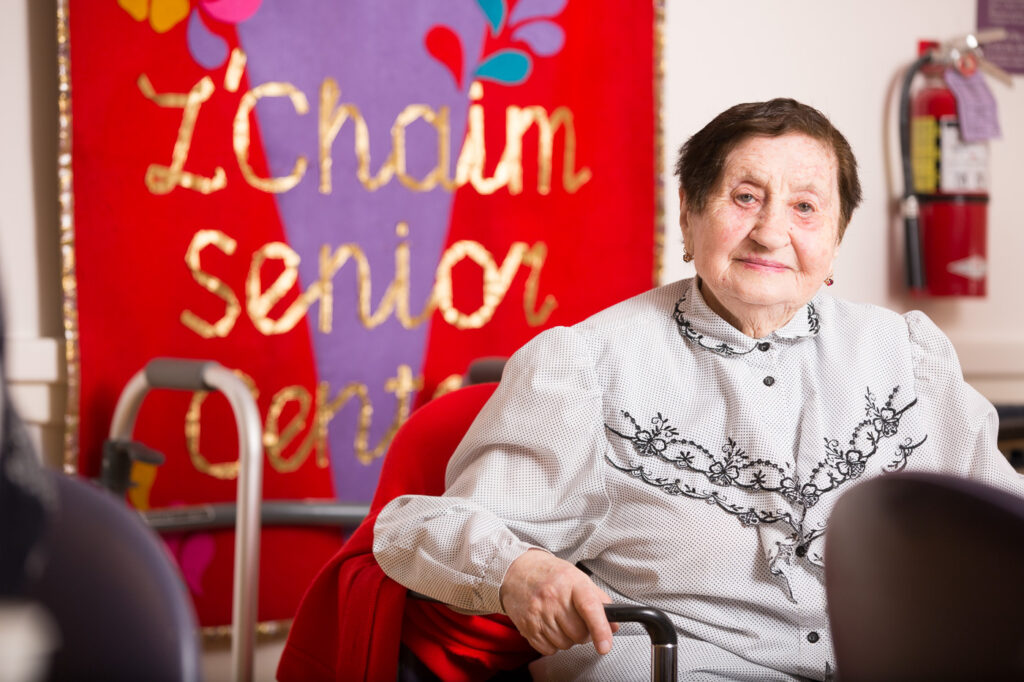 Older woman sitting in front of a L'Chaim Senior Center sign